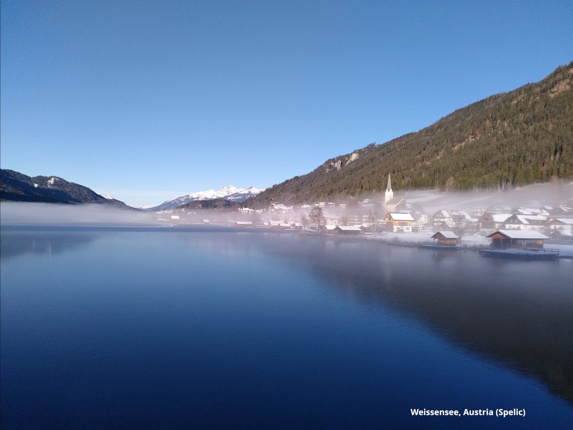 Mountain lake with light cloud strip across surface, houses on right in fog, greenish hills rise on right above, snow capped mountains far in the distance.