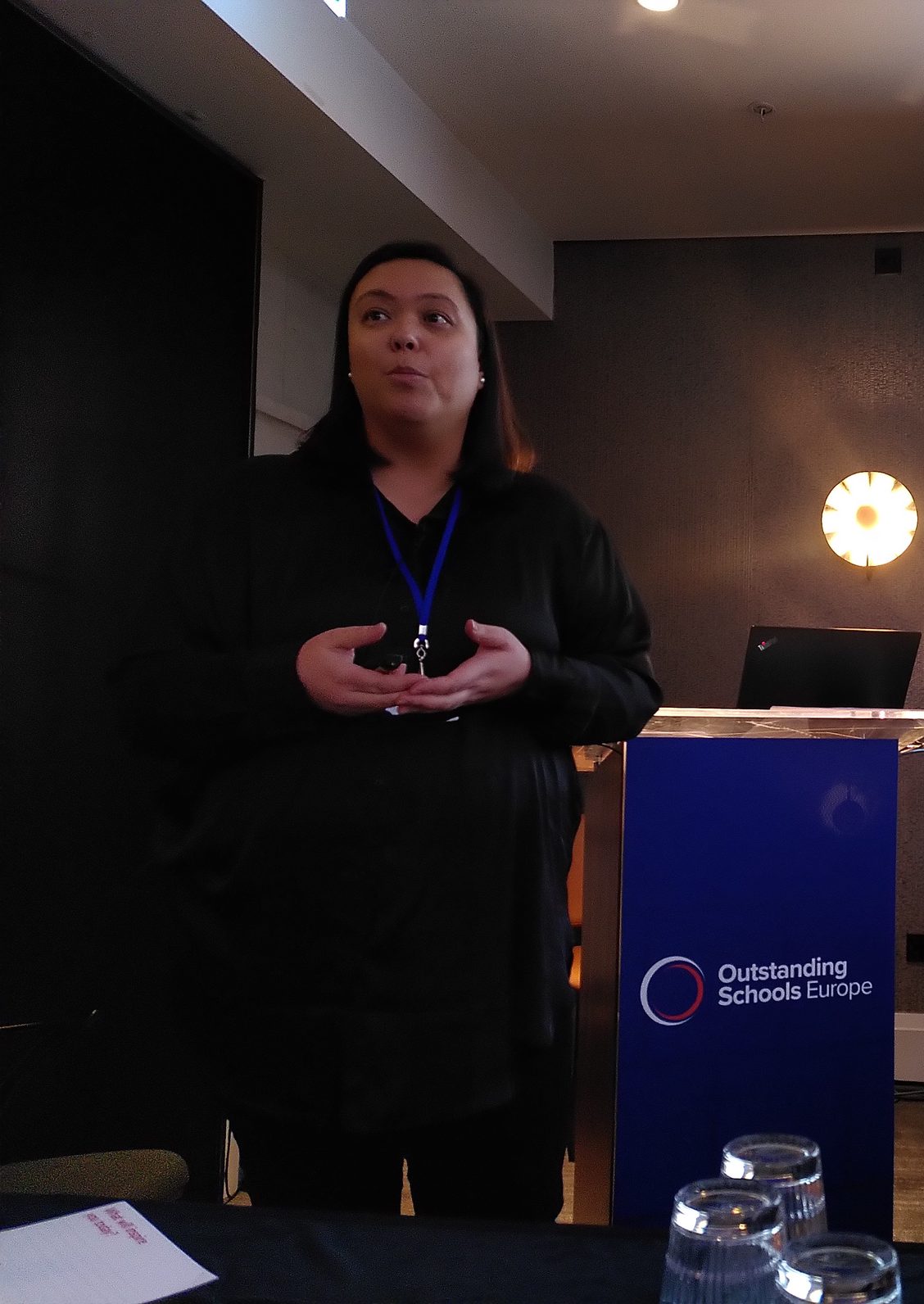 Australian-Filipino woman with straight shoulder length hair, dressed in black stands just in front and to the left of a lectern with Outstanding Schools Europe logo .